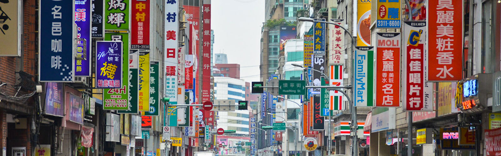 Brightly colored signs, covered in writing, hanging vertically off the sides of tall buildings along a street.